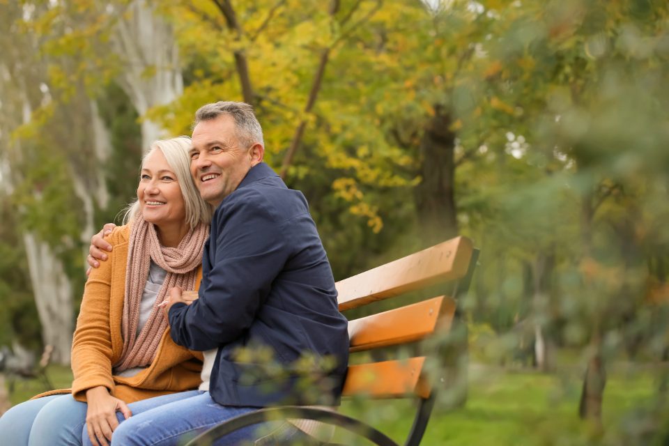 A smiling older couple sitting together on a park bench in autumn